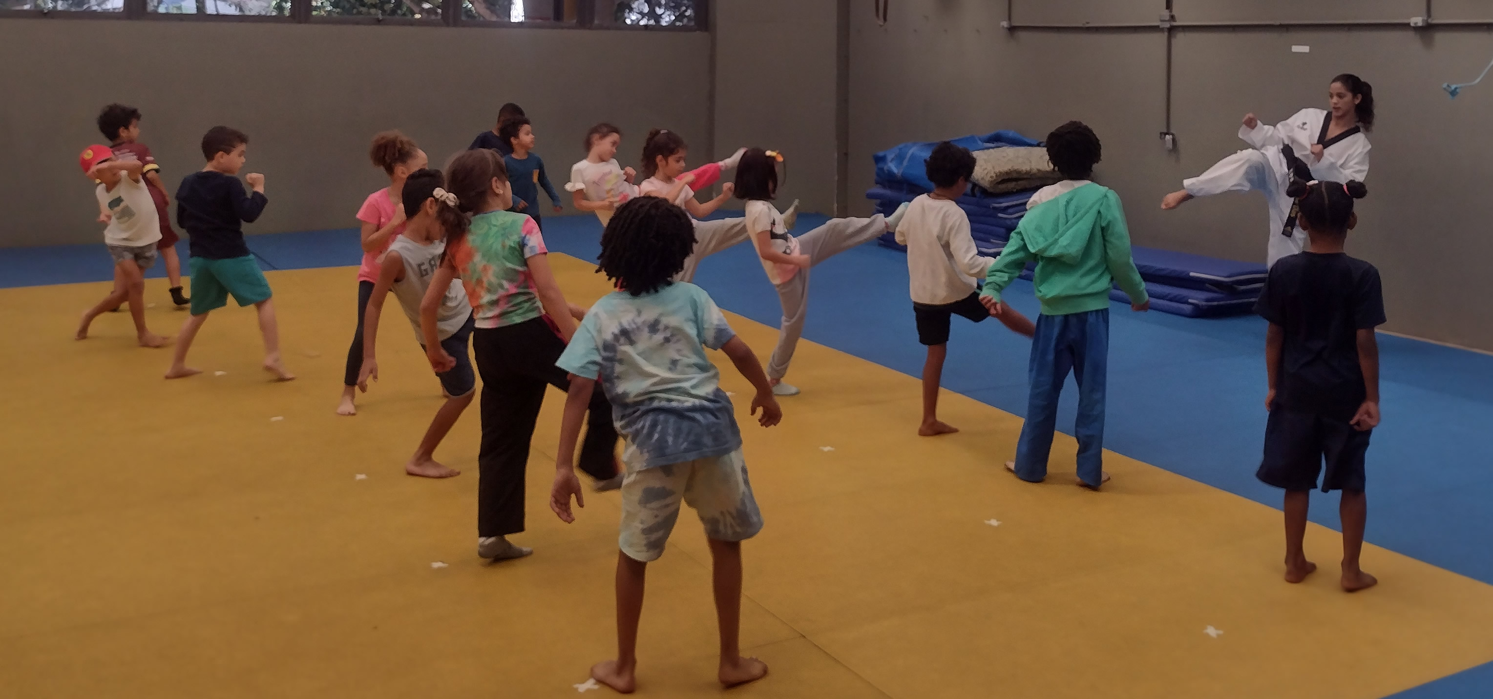Turma de iniciação infantil de Taekwondo com crianças em posição de chute, orientadas por instrutora faixa preta.